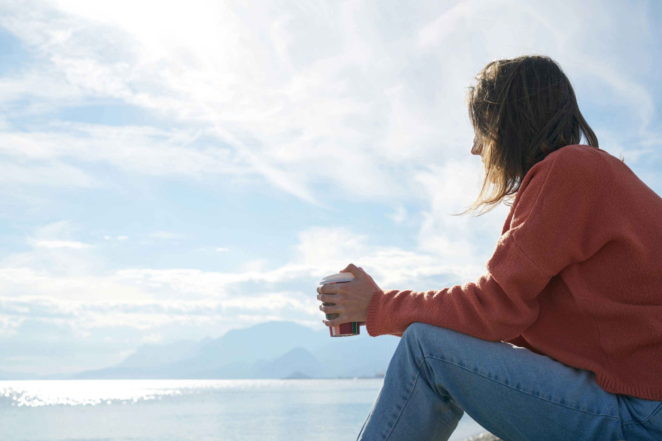 A person sitting beside a large body of water holding a coffee cup. 